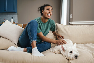 Happy black woman stroking her dog while resting on sofa at home