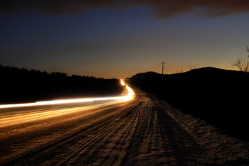 An asphalt road at dusk with the headlights of passing cars at a slow shutter speed. Night view...
