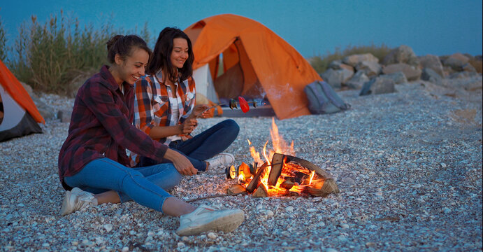 esbian couple roasting vegetables on fire, sitting on beach next to tent in camping.