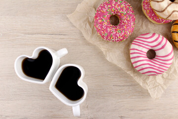 Two small white heart shaped mugs with coffee and donuts on wooden background