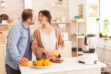 Obraz na płótnie Canvas Couple making smoothie in kitchen . Woman peeling of orange while smiling at husband. Healthy carefree and cheerful lifestyle, eating diet and preparing breakfast in cozy sunny morning