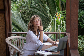 Portrait of young beautiful woman freelancer working with notebook, laptop in white shirt on balcony of tropical bungalow with palm trees view