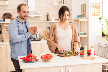 Obraz na płótnie Canvas Woman cutting cucumbers for healthy salad in kitchen while having a conversation with husband. Happy in love cheerful and carefree couple helping each other to prepare meal