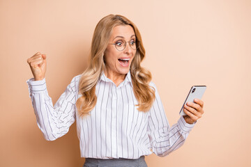 Fototapeta premium Photo of charming lucky mature woman dressed shirt glasses typing modern gadget rising fist isolated beige color background