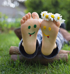 smiling faces are painted on the bare feet of a child lying on the green grass on a sunny day....