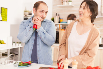 Husband looking at wife while she is smiling because of him in kitchen. Cutting red pepper on wooden chopper. Funny happy in love couple at home spending time together, cooking healthy and smiling