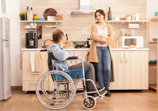 Disabled man in wheelchair spending time with wife in kitchen. Disabled paralyzed handicapped man with walking disability integrating after an accident.