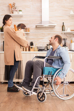 Wife holding fresh vegetables in paper bag after disabled husband in wheelchair doing groceries at supermarket. Disabled paralyzed handicapped man with walking disability integrating after an accident