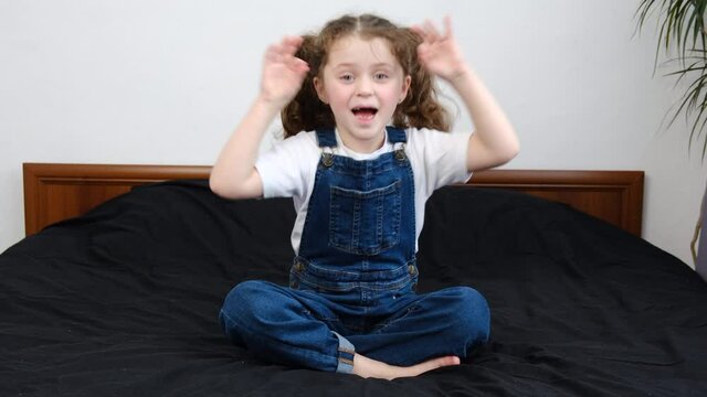 Smiling funny little girl kid sitting on bed at home play foolish show funny faces gesturing, playful cute small child grimace with tongue and hands, having fun looking at camera. Fools day concept