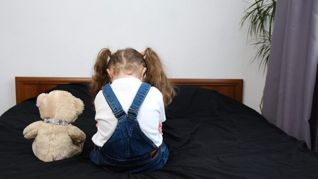 Back view of upset lonely bullied little kid girl sit on bed near teddy bear looking away feels abandoned abused, unhappy preschool child with thoughtful face, melancholic or bored. Autism concept
