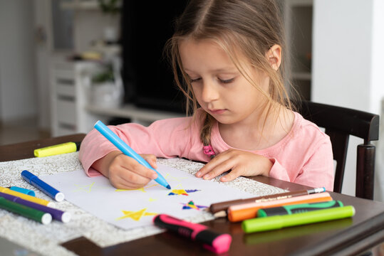 Little girl in pink t-shirt sits at table and draws stars. Kid is happy, she has lot of colored markers. Love for creativity, drawing, coloring pictures. Family, education of children.
