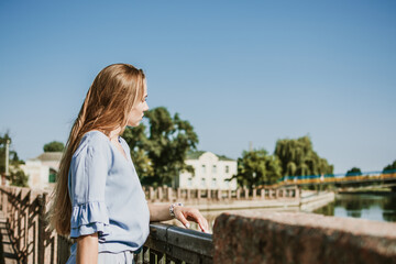 Fototapeta na wymiar Lonely pensive Young blonde woman in blue dress standing near the city river in summer sun day. Alone deep in thought woman outdoors