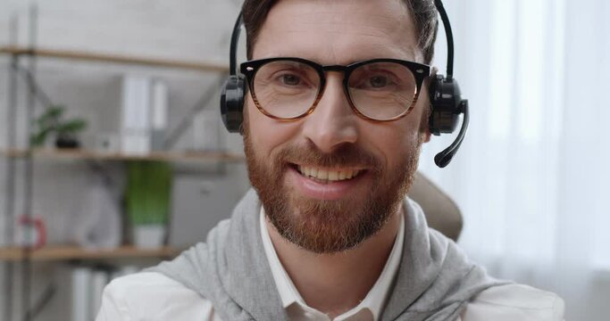 Portrait of Smiling Man with Eyeglasses and Headset looking at the Camera. Attractive Caucasian Bearded Man looking Successful sitting in Office Room. Working from Home. Having a Healthy Smile.