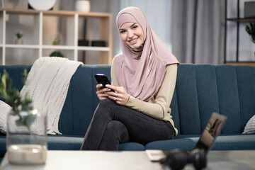Obraz premium Smiling muslim girl in beige headscarf using smartphone at home, typing message or reading news, while relaxing on comfortable blue sofa in living room.