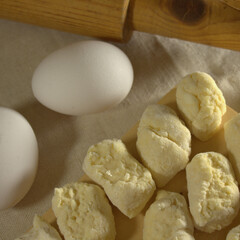 The process of making dumplings in the home kitchen. Pieces of raw dough are placed on a wooden board on linen cloth. Nearby is an old wooden rolling pin and a couple of chicken eggs.