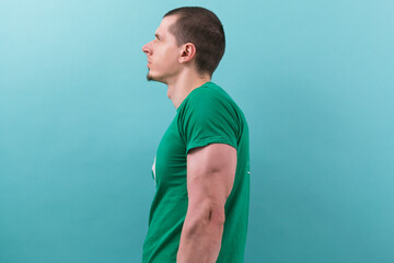 Caucasian man in green t-shirt with small black beard big arm stands side on a light blue...