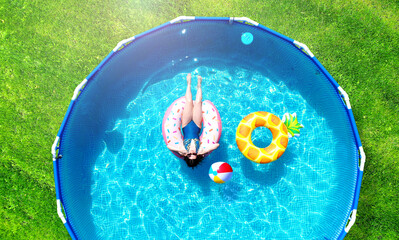 Aerial. Girl resting in a metal frame pool with inflatable toys. Summer leisure and fun concept. Frame pool stand on a green grass lawn. Top view from drone. - obrazy, fototapety, plakaty