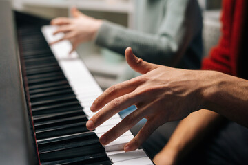 Close-up of unrecognizable child boy playing piano, music teacher sits near and helps with playing during lesson at music school. Kid practicing piano lesson in living room. Concept of music education