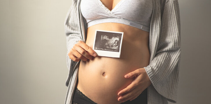 Pregnant woman holding an ultrasound picture next to her pregnant belly. The first photo of the baby. Maternal prenatal care. Maternity, pregnancy, baby expectation concept. Copy space