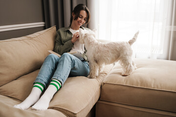 Happy white woman reading book while resting with her dog on sofa
