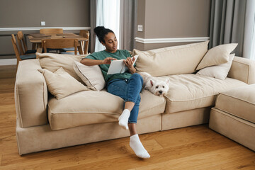 Obraz premium Smiling woman reading book while sitting with her dog on sofa at home