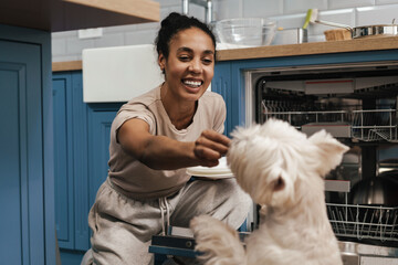Smiling black woman feeding her dog while using dishwasher