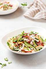 Rice noodle salad with cucumber, apple, onion, sesame seeds, and herbs in a white bowl on a light concrete background. Vegan salad Asian style. Vegetarian food. Vegetable salad with rice noodles
