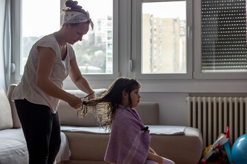 Young mother cutting her daughters hair at home during lockdown. Photo of cute girl with long blond hair having haircut at home. Mum doing haircut to her daughter at home during coronavirus outbreak.