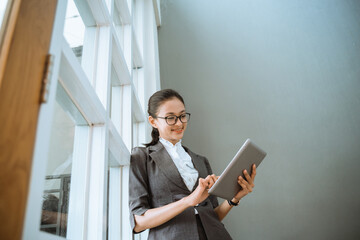 Portrait of young beautiful businesswoman looking at tablet standing and lean back in window office