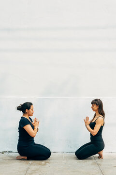 Vertical image of two women meditating on their knees facing each other dressed in black on the sidewalk of the street and in white background. Copy space