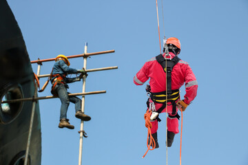 Working at height. A rear view of abseilers wearing red coverall and Personal Protective Equipment (PPE) in action hanging via rope access technique for touch up scaffolding activities.