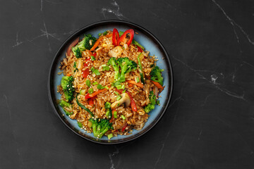 Asian rice with vegetables: zucchini, broccoli, red bel pepper, mushrooms, carrot, spring onion and sesame seeds. Dish isolated in a blue bowl, close-up on a black marble background. Asian cuisine.
