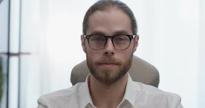 Portrait of Handsome Bearded Man in Eyeglasses Looking at the Camera at Office. Attractive Businessman Looking Successful while Sitting at Office Home Desk. Working Remotely. Distance. Smart People.
