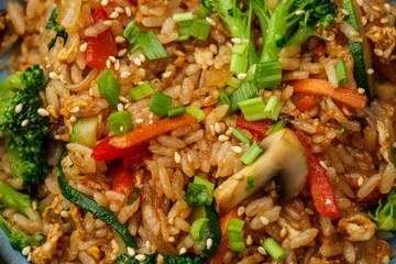 Asian rice with vegetables: zucchini, broccoli, red bel pepper, mushrooms, carrot, spring onion and sesame seeds. Dish isolated in a blue bowl, close-up on a black marble background. Asian cuisine.
