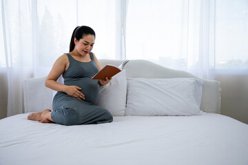 Happy young Asia pregnant woman holding and reading red book and sitting on white bed with copy space. Relaxation give birth mother
