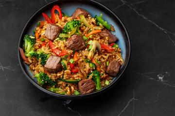 Asian rice with teriyaki beef and vegetables: zucchini, broccoli, pepper, mushrooms, carrot, onion and sesame seeds. Dish isolated in a blue bowl, close-up on a black marble background. Asian cuisine.