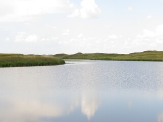 Small ripples on the water surface of the small lake.Cloudy warm summer day with a little breeze. Reflection of clouds in the water. Grass on the shore