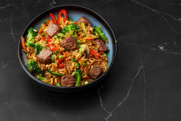 Asian rice with teriyaki beef and vegetables: zucchini, broccoli, pepper, mushrooms, carrot, onion and sesame seeds. Dish isolated in a blue bowl, close-up on a black marble background. Asian cuisine.