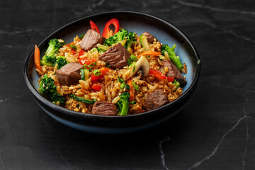 Asian rice with teriyaki beef and vegetables: zucchini, broccoli, pepper, mushrooms, carrot, onion and sesame seeds. Dish isolated in a blue bowl, close-up on a black marble background. Asian cuisine.
