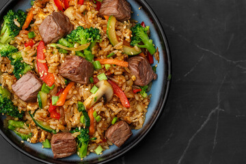 Asian rice with teriyaki beef and vegetables: zucchini, broccoli, pepper, mushrooms, carrot, onion and sesame seeds. Dish isolated in a blue bowl, close-up on a black marble background. Asian cuisine.