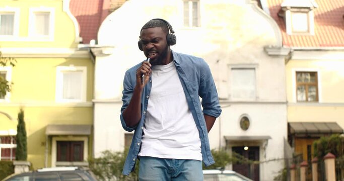 Cheerful Afro American Guy with Headphones Singing favourite Song in Smartphone. Having Positive Emotions Outdoors while Walking. Attractive Man listening to Music imagines himself on stage. Fun.
