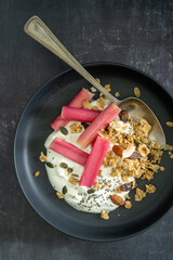 Close up from above of yoghurt and rhubarb with granola in a black bowl and a pan with rhubarb on black background, with copy space