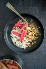 Flat lay of yoghurt and rhubarb with granola in a black bowl and a pan with rhubarb on black background, with copy space