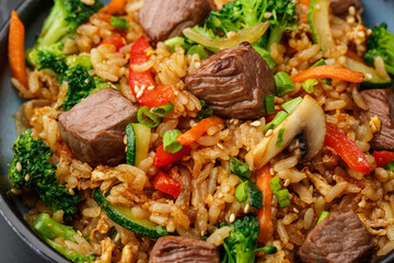 Asian rice with teriyaki beef and vegetables: zucchini, broccoli, pepper, mushrooms, carrot, onion and sesame seeds. Dish isolated in a blue bowl, close-up on a black marble background. Asian cuisine.