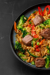 Asian rice with teriyaki beef and vegetables: zucchini, broccoli, pepper, mushrooms, carrot, onion and sesame seeds. Dish isolated in a blue bowl, close-up on a black marble background. Asian cuisine.