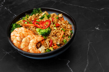 Asian rice with shrimps and vegetables: zucchini, broccoli, pepper, mushrooms, carrot, onion and sesame seeds. Dish isolated in a blue bowl, close-up on a black marble background. Asian cuisine.