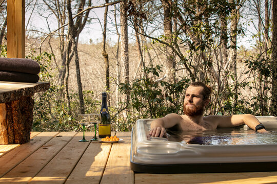 Young hipster man lounging in a jacuzzi in the woods with a bottle of champagne. 