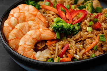 Asian rice with shrimps and vegetables: zucchini, broccoli, pepper, mushrooms, carrot, onion and sesame seeds. Dish isolated in a blue bowl, close-up on a black marble background. Asian cuisine.