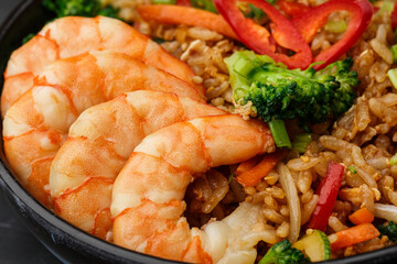 Asian rice with shrimps and vegetables: zucchini, broccoli, pepper, mushrooms, carrot, onion and sesame seeds. Dish isolated in a blue bowl, close-up on a black marble background. Asian cuisine.