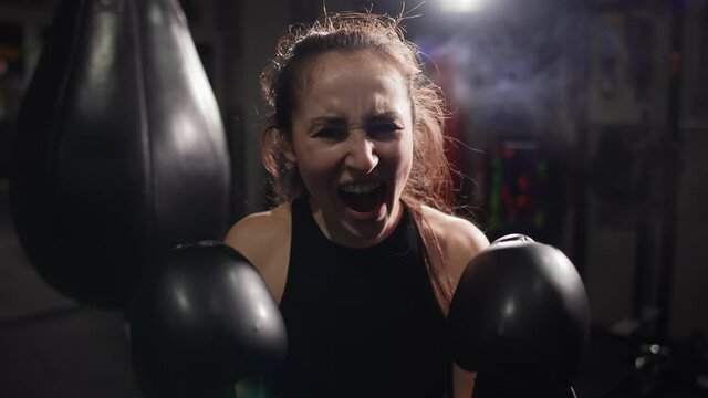 Female boxer standing at dark gym, looking intensely at the camera and screaming